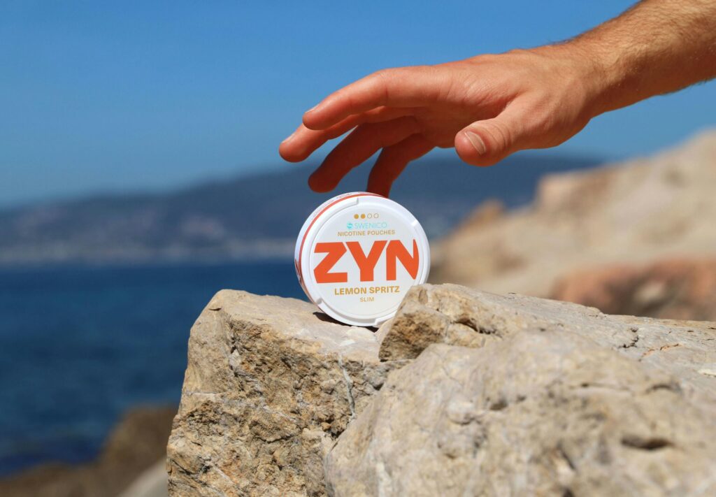 Close-up of a hand reaching for a ZYN nicotine pouch on a rock by the sea.
